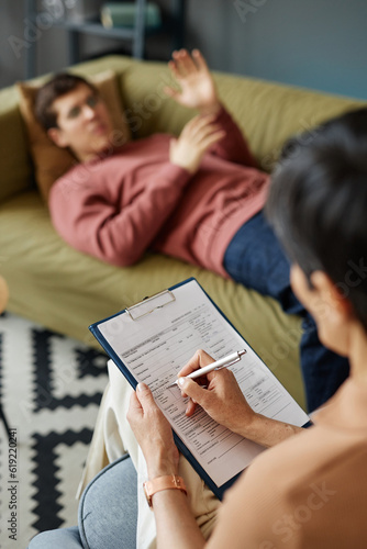 Vertical image of psychologist filling card of patient while he lying on sofa and telling about his problems