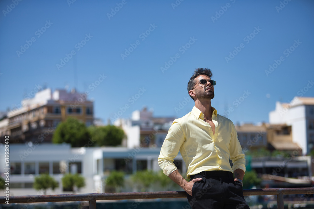 Young and attractive businessman, with beard, yellow shirt, black pants and sunglasses, with his hands in his pockets and looking at the sky leaning on a railing. Concept beauty, fashion, success.