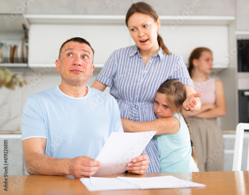 Angry woman shouts at husband for negligent use of family budget  he calculates  reads report  counts interest on loan. Youngest daughter hugs and supports mother in family conflict
