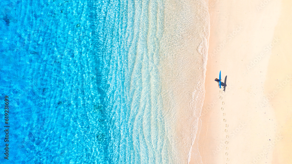 Surfer with a board on the beach. Blue water and clean beach in the summer. View from a drone. An aerial view of the seascape. Recreation and active sports.