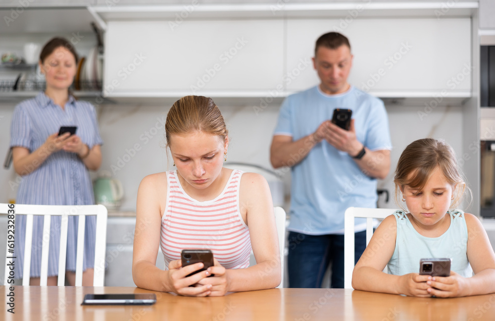 Fototapeta premium Sister girls sitting near kitchen table with phones in hands and flipping through tape and liking photos of friends, parents surf Internet in background