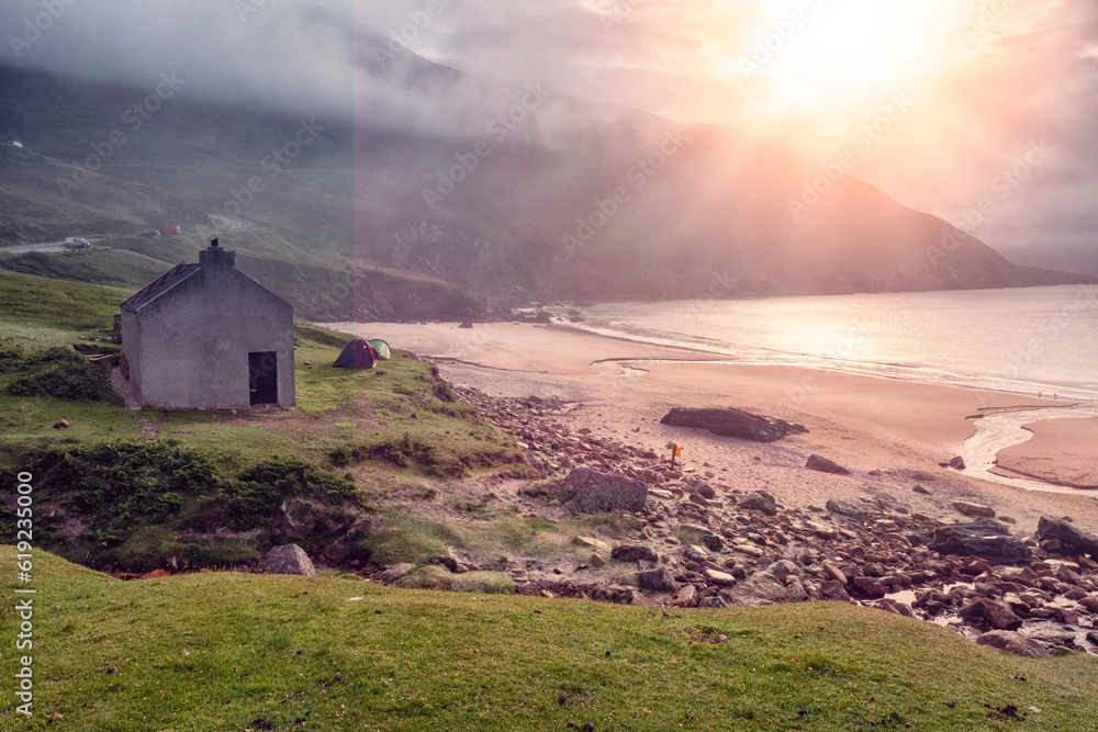 Small house by the ocean and beach, sun rise over a cliff and low cloudy sky. Keem beach, Ireland. Popular travel area, Irish nature scene. Calm mood. Small house by the ocean and beach, sun rise over a cliff and low cloudy sky. Keem beach, Ireland. Popular travel area, Irish nature scene. Calm mood.