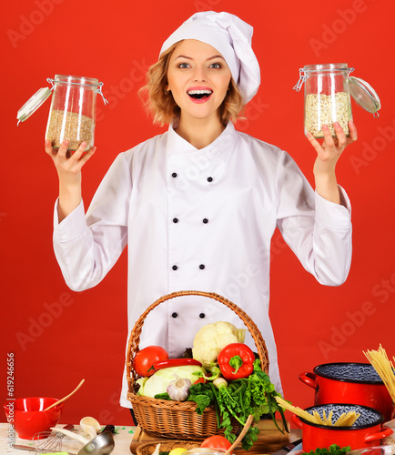 Smiling chef in white uniform standing at table with basket vegetables holds glass jars with groats. Healthy food cooking. Female cook holds jars with porridge for breakfast. Preparation organic food. photo