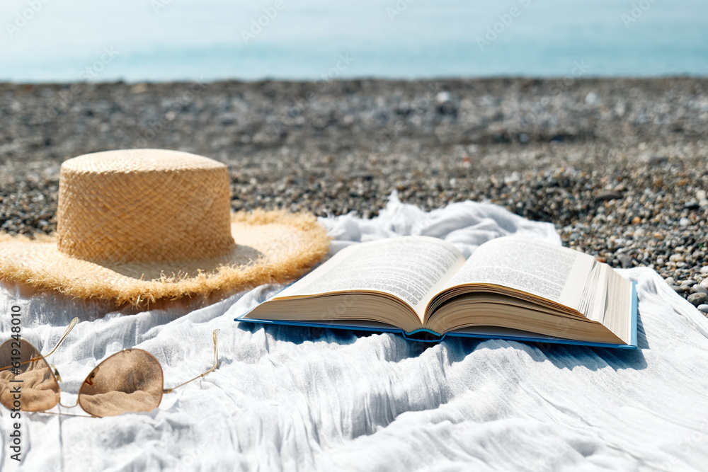 Open book, sunglasses and straw hat on the towel on pebble beach. Concept of reading and relaxing in summer vacation. Beach literature. Open book, sunglasses and straw hat on the towel on pebble beach. Concept of reading and relaxing in summer vacation. Beach literature.