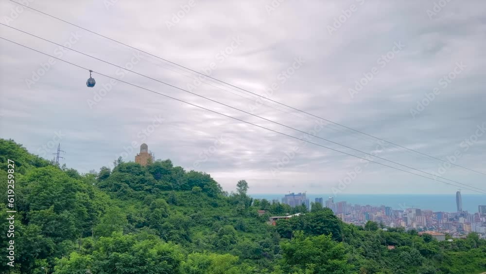 Ropeway, cable car cabins are moving against the cloudy gray sky in Batumi, Georgia - aerial rope transit, gondola lift. Georgian landscape with mountains, trees and monastery Ropeway, cable car cabins are moving against the cloudy gray sky in Batumi, Georgia - aerial rope transit, gondola lift. Georgian landscape with mountains, trees and monastery