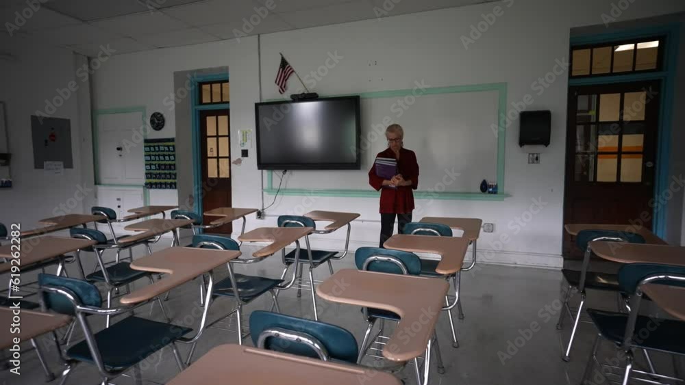 Wide angle push in view of sad teacher in an empty classroom with the lights turned off during a pandemic wondering about the future of education. Wide angle push in view of sad teacher in an empty classroom with the lights turned off during a pandemic wondering about the future of education.