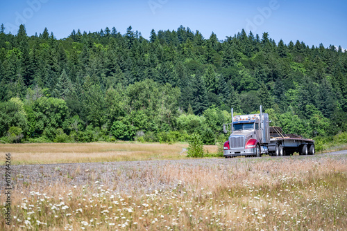 Two color bonnet big rig semi truck with lot of accessories transporting fastened lumber wood on the flat bed semi trailer driving on the scenic nature road
