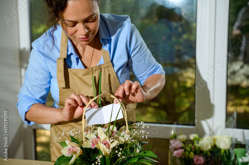Close-up female florist inserting a white empty congratulatory card over a cute flower arrangement in a wicker basket