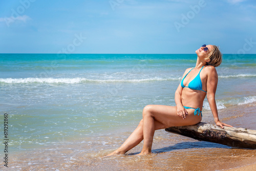 A young woman in a blue swimsuit and sunglasses sits on the seashore on a sunny day. Tourism  travel and vacation in the resort. Space for text.