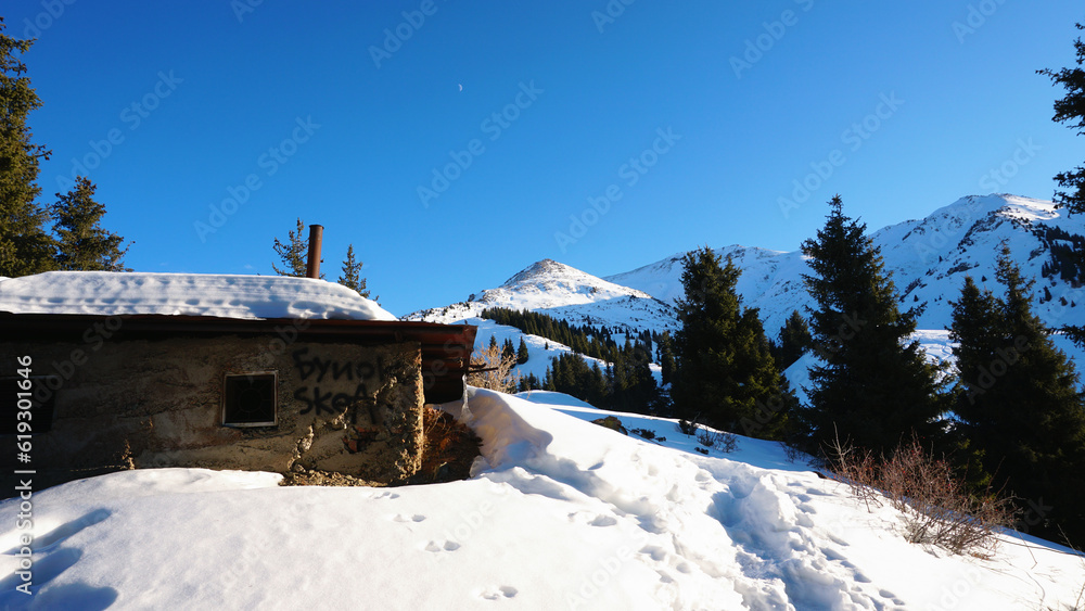 An abandoned house in the snowy mountains. Christmas trees and bushes grow. Clear sky and moon. A small moon in the distance above the top of the peak. There is a lot of snow on the roof. Almaty