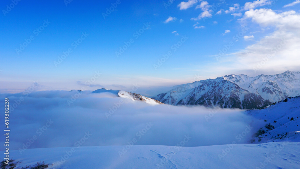 An ocean of clouds in the snowy mountains at dawn. White clouds are like a carpet in a gorge. Waves rise and descend from the mountains. Sunrise of the yellow sun. Clear sky, mountain peaks. Almaty An ocean of clouds in the snowy mountains at dawn. White clouds are like a carpet in a gorge. Waves rise and descend from the mountains. Sunrise of the yellow sun. Clear sky, mountain peaks. Almaty
