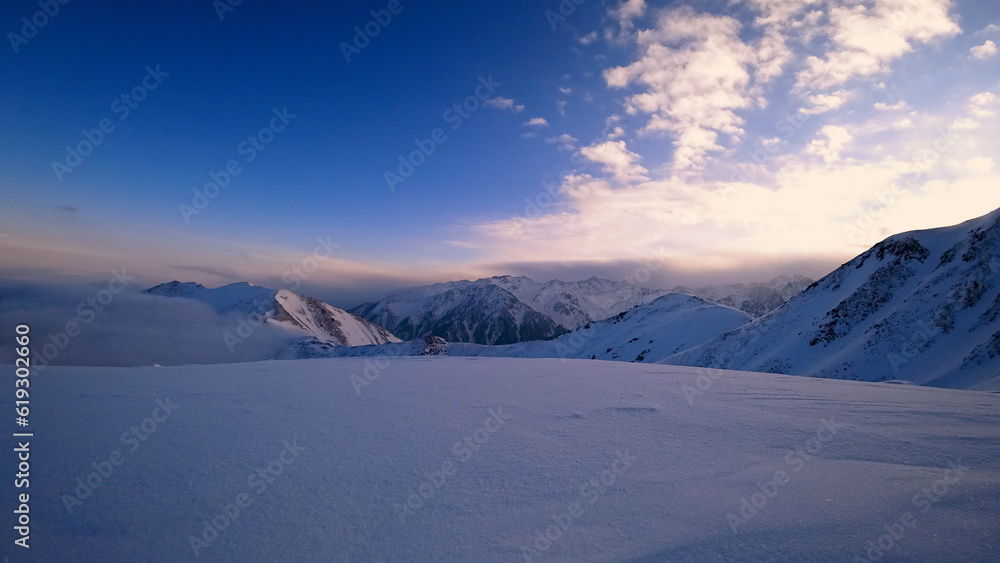 An ocean of clouds in the snowy mountains at dawn. White clouds are like a carpet in a gorge. Waves rise and descend from the mountains. Sunrise of the yellow sun. Clear sky, mountain peaks. Almaty