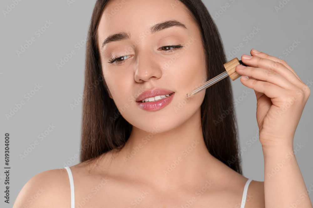 Beautiful young woman applying essential oil onto face on grey background Beautiful young woman applying essential oil onto face on grey background