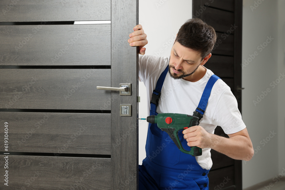 Worker in uniform with screw gun repairing door lock indoors