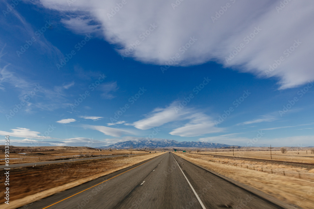 Landscape with a highway among meadows and trees in the foreground, blue mountains with snow in the background and a bright blue sky with large white clouds. Scenic landscape on a sunny autumn day Landscape with a highway among meadows and trees in the foreground, blue mountains with snow in the background and a bright blue sky with large white clouds. Scenic landscape on a sunny autumn day