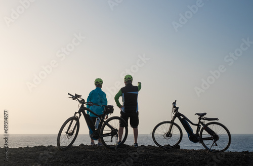 Back view of senior couple of cyclists standing face the sea with their bikes looking at the horizon over water. Elderly man and woman enjoying freedom and healthy lifestyle