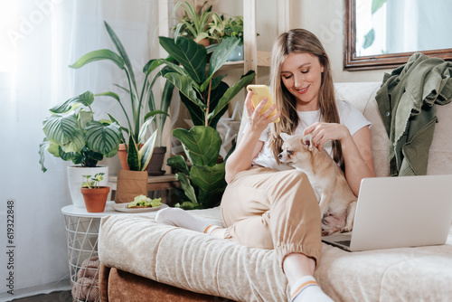 Freelance woman with dog and laptop, texting at mobile phone and working from home office. Happy girl sitting on couch in living room with plants. Distance learning online education and work.