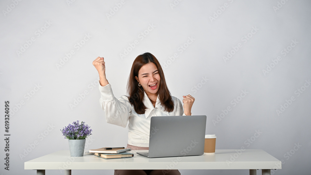 A cheerful Asian businesswoman sits at her desk, raising her hands, screaming with joy A cheerful Asian businesswoman sits at her desk, raising her hands, screaming with joy