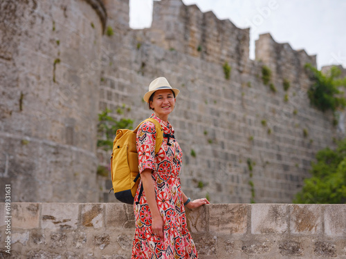 summer trip to Rhodes island, Greece. Young Asian woman in ethnic red dress walks over one of gates Rhodes fortress, road over moat. female traveler visiting southern Europe Unesco world heritage site