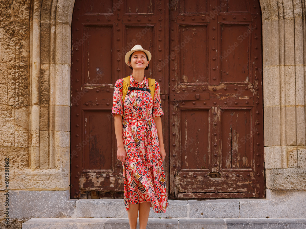 summer trip to Rhodes island, Greece. Young Asian woman in ethnic red dress stand near main entrance to Master's castle Rhodes. female traveler visiting southern Europe. Unesco world heritage site.