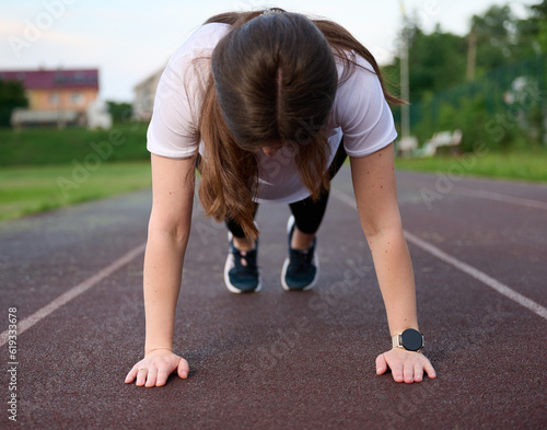 Сlose-up view of a pretty girl performing a plank on the jogging track. Workout. Young attractive female in sports tight clothes doing stretching exercises and warming up for jogging in the stadium.