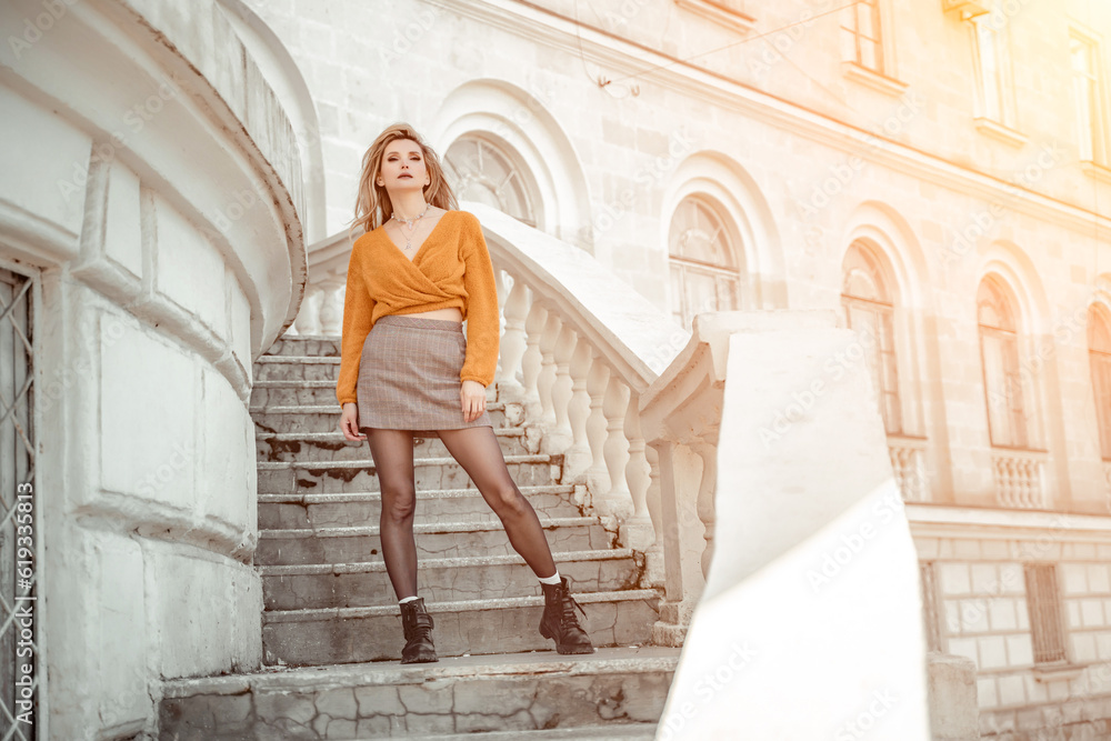 A middle-aged woman looks like a good blonde with curly beautiful hair and makeup on the background of the building. She is wearing a yellow sweater.
