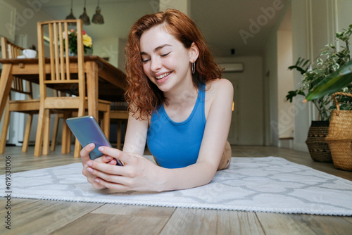 Happy young woman using smart phone on rug at home