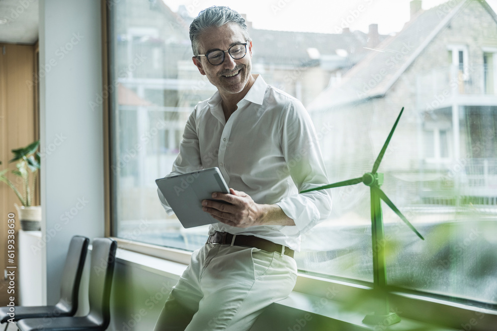 Smiling mature businessman with tablet PC looking at wind turbine model in office Smiling mature businessman with tablet PC looking at wind turbine model in office