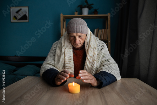 Fotografie Old man dressed in a warm winter clothes sits at home at a table and warms his hands from a burning candle