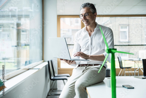 Smiling businessman sitting on desk using laptop by wind turbine model in office photo