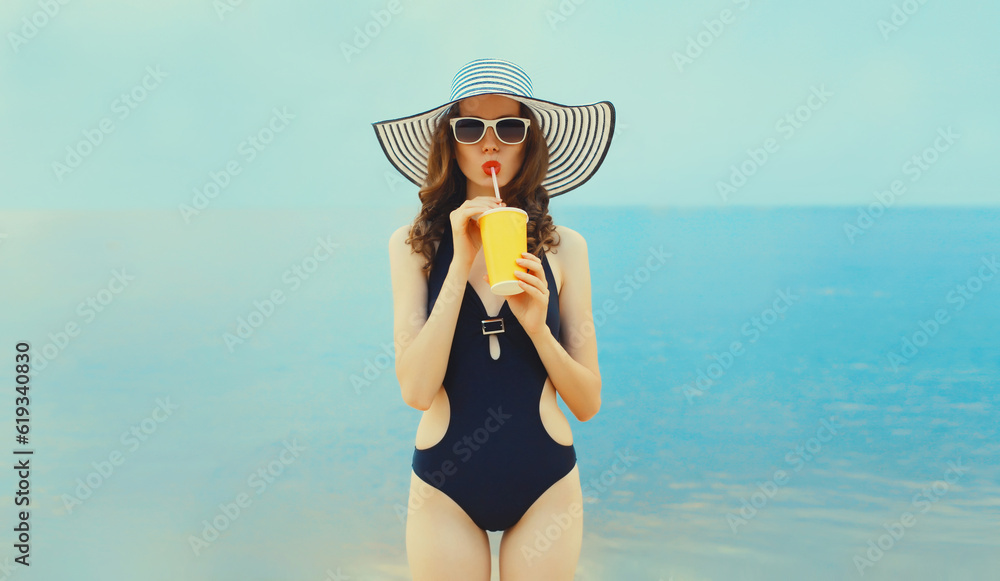 Summer portrait of young woman model drinking fresh juice wearing swimsuit, straw hat on the beach on sea background
