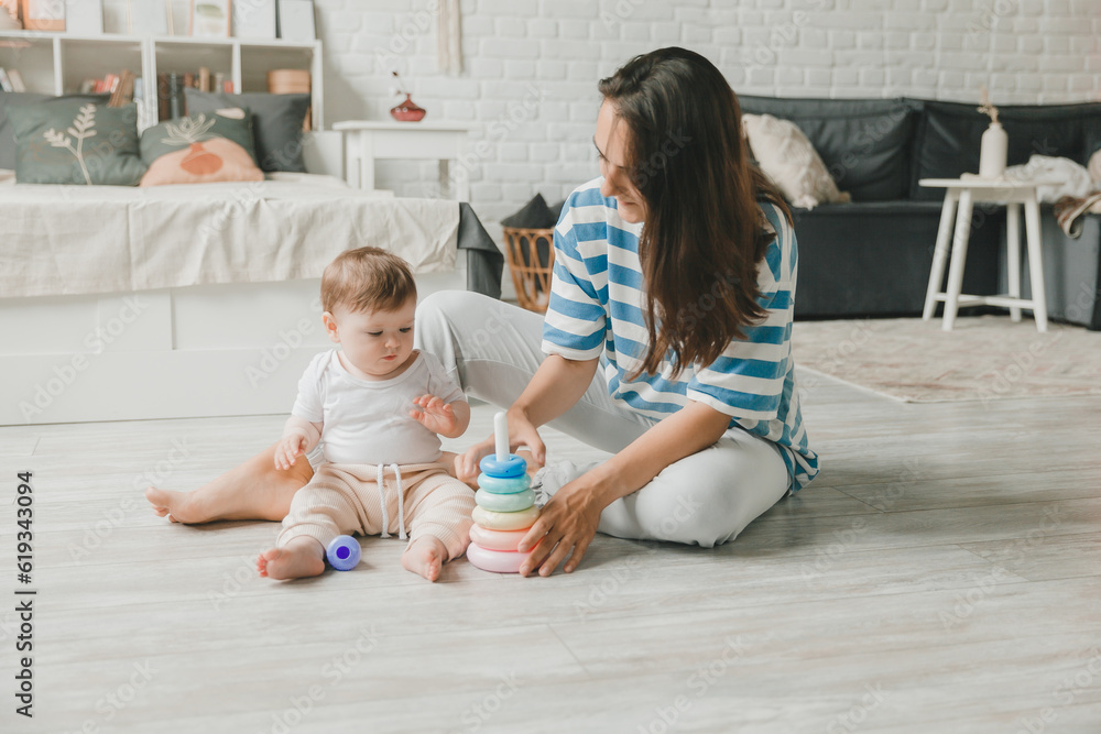 Fototapeta premium Beautiful young mother plays and teaches her baby 6 months old on the floor in the living room.mom and baby play with toys