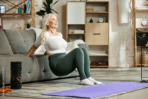 Smiling, beautiful, senior woman training at home in living room, doing exercises with couch. Strong fit body. Concept of sportive and healthy lifestyle, age, body care, fitness, wellness.