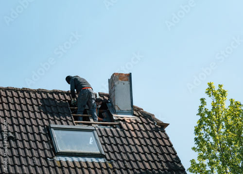 A roofer stands atop a house, destroying the chimney from an unusually low angle view. Its distinctive architecture and tiled roof stand out against the clear sky.