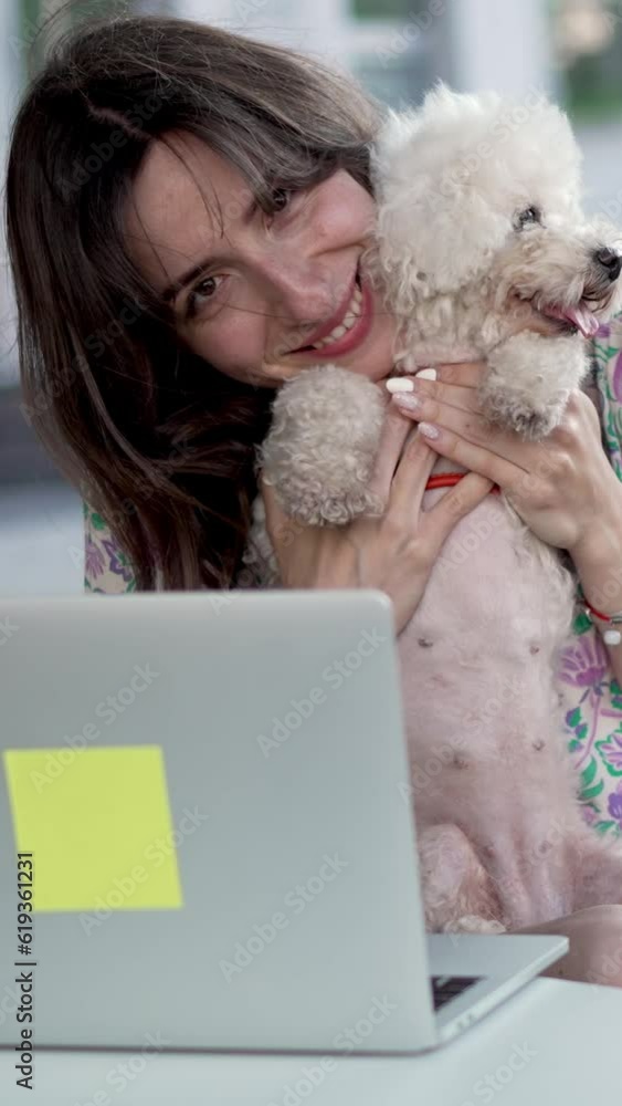 Slow motion of smiling young woman with long dark hair playing with Bichon Frise dog sitting at table with laptop at sidewalk city cafe Vertical video Slow motion of smiling young woman with long dark hair playing with Bichon Frise dog sitting at table with laptop at sidewalk city cafe Vertical video