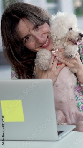 Slow motion of smiling young woman with long dark hair playing with Bichon Frise dog sitting at table with laptop at sidewalk city cafe Vertical video