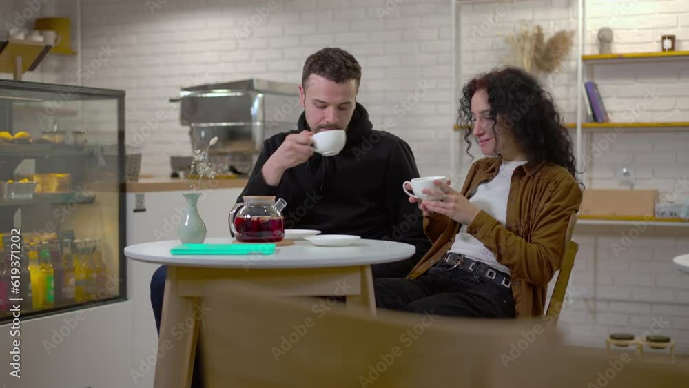Young Caucasian man and woman drinking tea talking dating in cafe in urban city. Positive relaxed boyfriend and girlfriend enjoying date in coffee house Young Caucasian man and woman drinking tea talking dating in cafe in urban city. Positive relaxed boyfriend and girlfriend enjoying date in coffee house