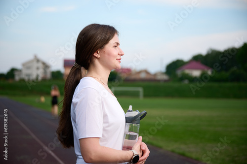 Portrait of the slim fit young girl with bottle of water. Sporty female standing near the stadium on the jogging track. photo