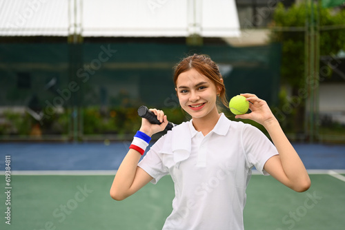 Cheerful sportswoman holding racket and ball standing on the tennis court. Fitness, sport and competition concept © Prathankarnpap