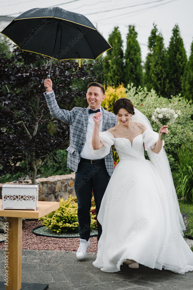 Happy bride and groom in rain with an umbrella outdoors. Newlyweds with bouquet standing on wedding ceremony in backyard banquet area. Fidelity in love. Couple says promise, exchanging vows in nature.