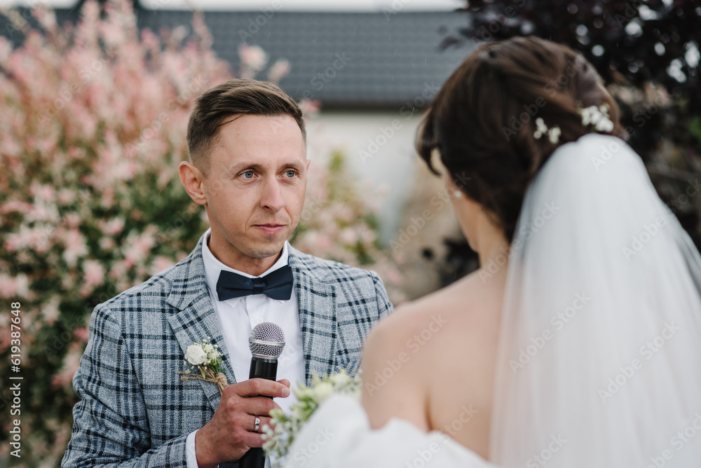 Groom says the promise in the microphone in nature. Happy bride and groom exchanging wedding vows on ceremony in the backyard banquet area. Newlyweds standing with bouquet. Fidelity in love. Closeup. Groom says the promise in the microphone in nature. Happy bride and groom exchanging wedding vows on ceremony in the backyard banquet area. Newlyweds standing with bouquet. Fidelity in love. Closeup.