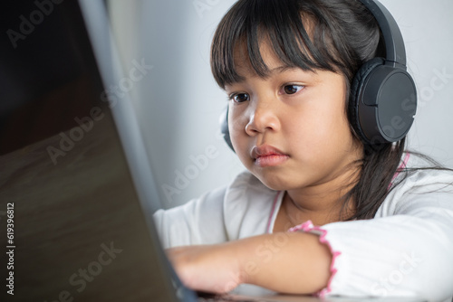 4-5 years old Asian girl wearing white shirt is studying online via computer using bluetooth headphones at home, distance education concept