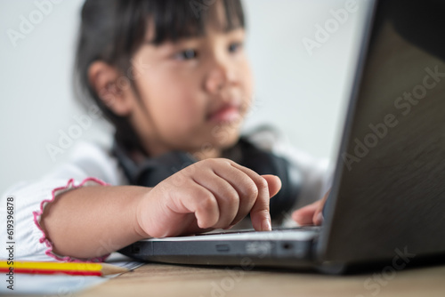 5-6 years old Asian girl wearing white shirt is studying online via computer using bluetooth headphones at home, distance education concept