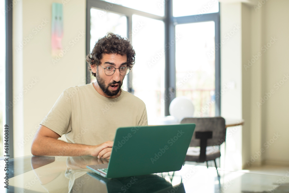 young adult bearded man with a laptop looking very shocked or surprised, staring with open mouth saying wow young adult bearded man with a laptop looking very shocked or surprised, staring with open mouth saying wow