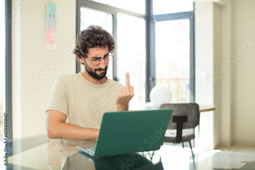 young adult bearded man with a laptop feeling angry, annoyed, rebellious and aggressive, flipping the middle finger, fighting back