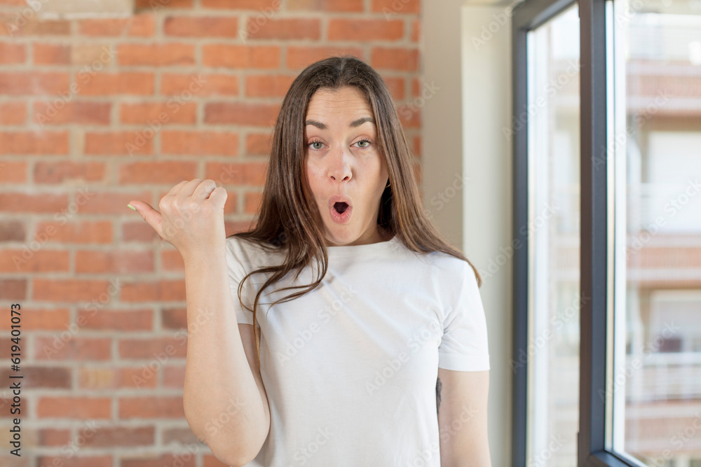 young adult pretty woman looking astonished in disbelief, pointing at object on the side and saying wow, unbelievable