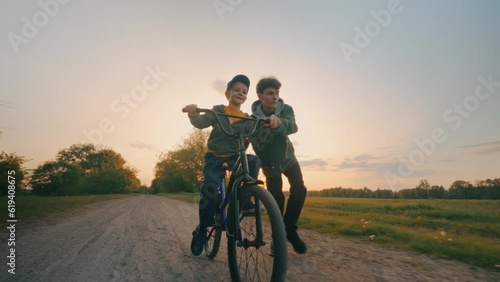 The big brother teaches the younger to ride a bike on a rural road at sunset.