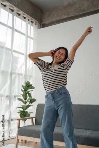 Fototapeta Naklejka Na Ścianę i Meble -  Attractive happy young Asian woman listening music with headphones from music application dance raise up hand near grey sofa couch stay at home rest relax spend free spare time in living room indoor