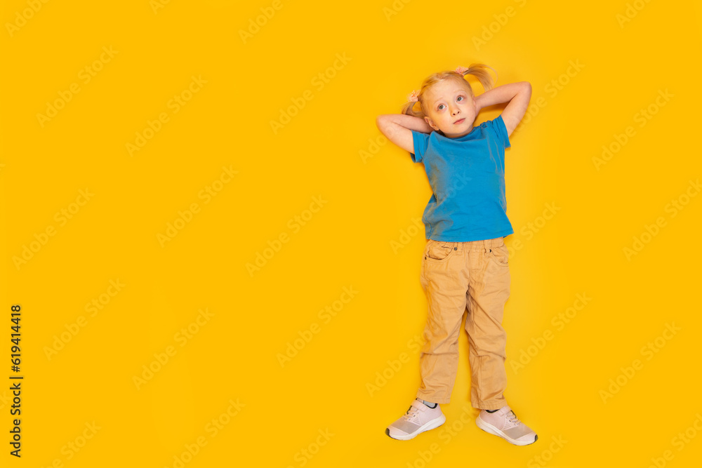 Child lying on yellow background. Little girl with two ponytails wears blue T-shirt and yellow pants. Top view. Copy space, mockup Child lying on yellow background. Little girl with two ponytails wears blue T-shirt and yellow pants. Top view. Copy space, mockup
