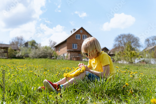 Child rests countryside during summer holidays. Blond boy sits on the grass and weaves wreath from dandelions photo